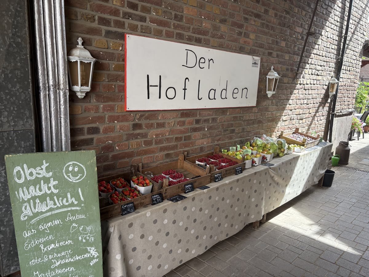 Handgeschriebenes Schild „Der Hofladen“ an der Backsteinwand, darunter die lange Theke mit Obst und eine grüne Tafel mit der Aufschrift „Obst macht glücklich“