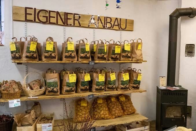 Holzregal unter dem Schild „Eigener Anbau“ mit Kartoffeln in Papiertüten und Körben